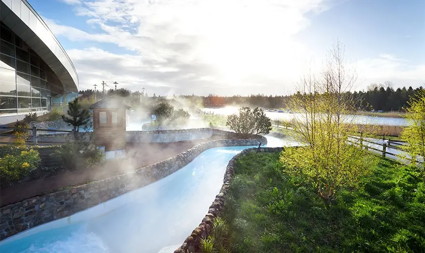 Outdoor spa with warm water stream, surrounded by lush greenery and mist, under a bright sky near a modern building.